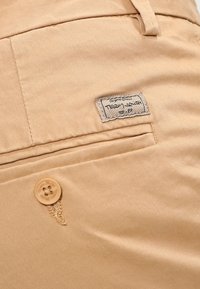 Gros plan sur la poche d'un pantalon beige avec une fermeture à bouton et une étiquette en tissu cousue portant l'inscription "TEDDY_SMITH 75-89".