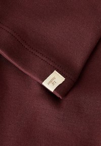 Gros plan sur un tissu bourgogne avec des détails de couture et une petite étiquette beige portant la mention "Lif Algeier."