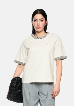 Woman with straight black hair wears a white short-sleeve top with patterned trim and holds a black puffy handbag at her side.