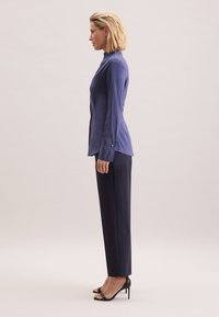 Donkereblauwe blouse met lange mouwen, een hoge kraag en knopen aan de zijkant, gecombineerd met op maat gemaakte donkere broek en zwarte open schoenen met hakken.