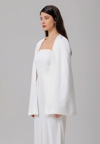 Une cape en satin blanc avec une silhouette structurée, un design minimaliste et une texture lisse, complétant une robe sans manches ajustée.