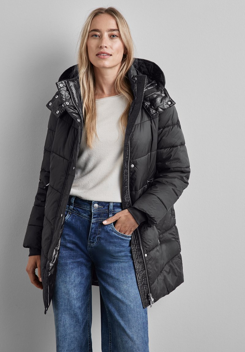 Svart puffjacka med huva, med en kviltad design och glänsande detaljer. Kombinerad med ljusblå vida jeans och en beige topp.