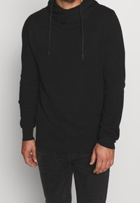 Černá pletená mikina s kapucí s límcem na stažení, texturovaným materiálem a šňůrkami na stažení. Má dlouhé rukávy a volný střih.