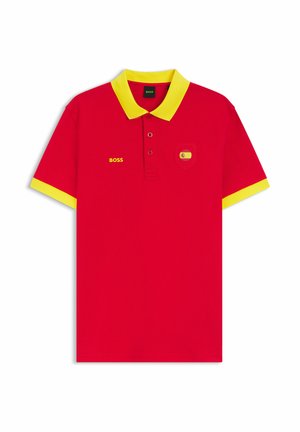 Czerwona koszulka polo z żółtym kołnierzem i mankietami, napis "BOSS" na lewej stronie klatki piersiowej oraz mała naszywka z flagą Hiszpanii na prawym kieszonce na klatce piersiowej.