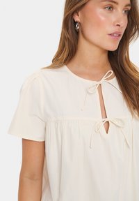 Camicia beige chiara con maniche corte, scollatura rotonda e apertura a chiave legata con un fiocco. Tessuto liscio con leggero volume all'orlo.