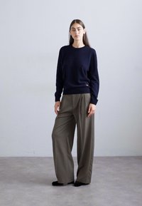 Woman standing wearing a navy long-sleeve sweater and loose olive-green wide-leg trousers against a plain light gray background.