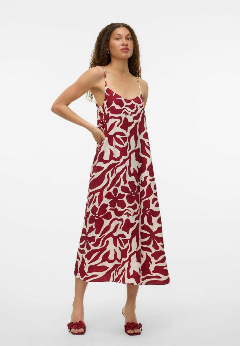 Robe maxi crème avec des motifs floraux abstraits rouges audacieux, dotée de fines bretelles et d'une coupe décontractée. Portée avec des sandales à glisser assorties.
