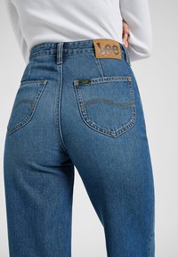 Jeans in denim blu con vita alta, cuciture visibili, due tasche posteriori e un'etichetta in pelle marrone con il marchio "Lee".