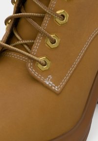 Primo piano di un boot in pelle color corda con spesse lacci infilati attraverso occhielli esagonali dorati e dettagli in cucitura bianca.