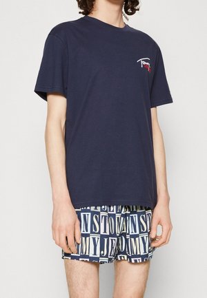 Młoda osoba w granatowej koszulce z małym logo i nadrukowanych szortach z pogrubionymi literami układającymi napis "Tommy Jeans."