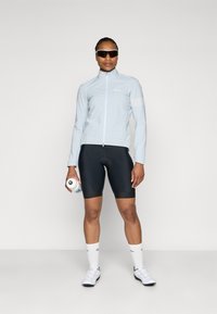 Light blue cycling jacket with a stand-up collar and zipper, paired with black cycling shorts. Also features white cycling shoes and a water bottle.