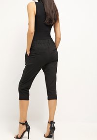 Pantalon noir raccourci à coupe décontractée, avec une taille élastique et des poches latérales. Porté avec des sandales à talons hauts noires.