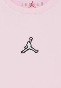 Vaaleanpunainen puuvillapaita, jossa on musta ja harmaa brodeerattu Jumpman-logo edessä. Pyöreä kaula-aukko ja sileä materiaali.