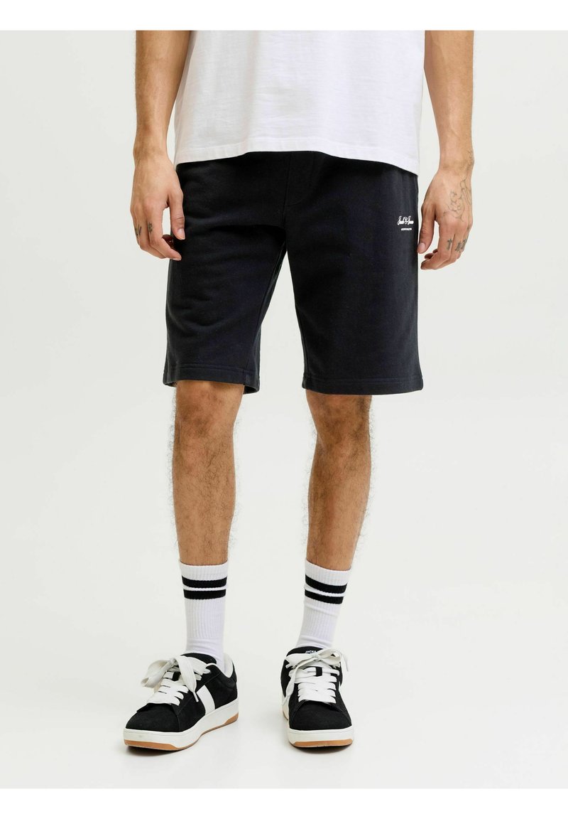 Homme portant un short noir, des chaussettes blanches à rayures noires, des baskets noires et blanches, et un t-shirt blanc sur un fond uni.