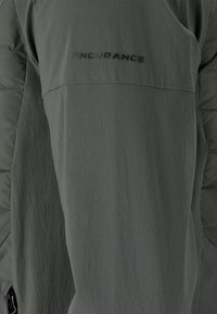 Grå ytterkläder med en texturerad tygkvalitet som har en lätt glans. Ärmen har en diskret svart etikett med texten "ENDURANCE."