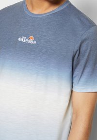 Personne portant un T-shirt à dégradé bleu vers blanc à manches courtes avec le logo ellesse brodé sur la poitrine.