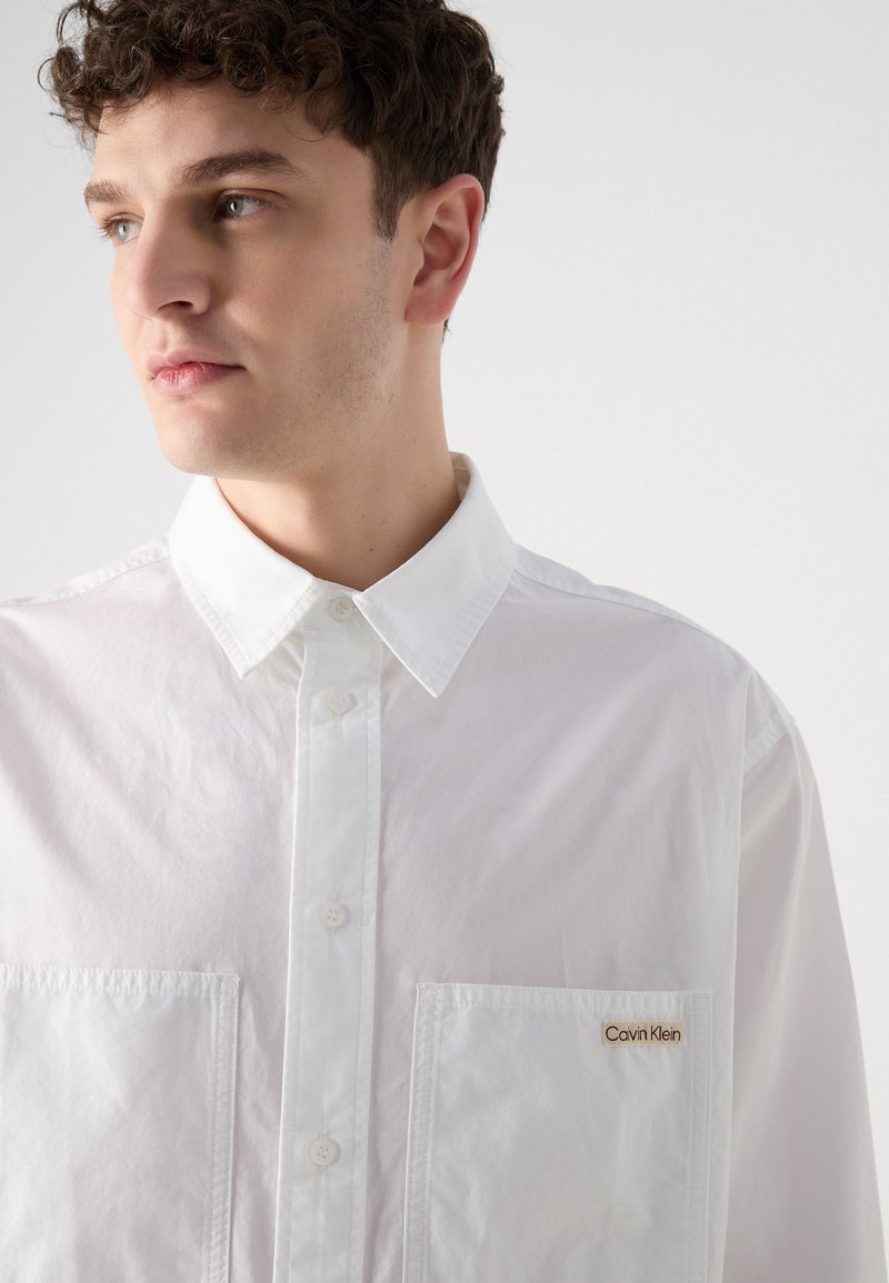 Camicia bianca con bottoni, dotata di un colletto classico, tasche frontali e un piccolo logo dorato sulla tasca sinistra. Tessuto leggero e liscio.