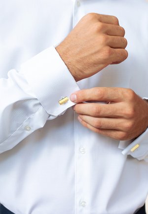 Weißes Hemd mit strukturierter Optik, ausgestattet mit goldenen Manschettenknöpfen. Die Manschette wird angepasst, während eine Knopf-Leiste sichtbar ist.