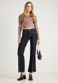 Temno modri kavbojasti hlače s širokimi hlačnicami, kontrastnimi šivi in stranskimi zadrgami. Usklajene z prosojno karo dolgo rukavno majico in sijočimi čevlji na peto.