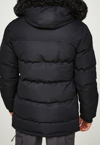 Chaqueta acolchada negra con capucha de piel de pelo desmontable, que presenta una textura acolchada y puños de canalé. Incluye un logotipo sutil en la manga.