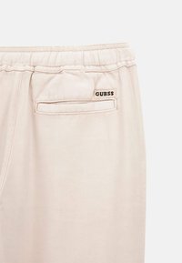 Beige katoenen broek met een elastische tailleband, voorzien van een achterzak met een witte rand en een zwart "GUESS" logo label.