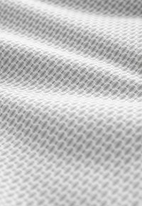 Fehér, texturált szövet kis gyémánt alakú geometriai mintával, amely puha, réteges megjelenést kölcsönöz, látható hullámokkal és redőkkel.