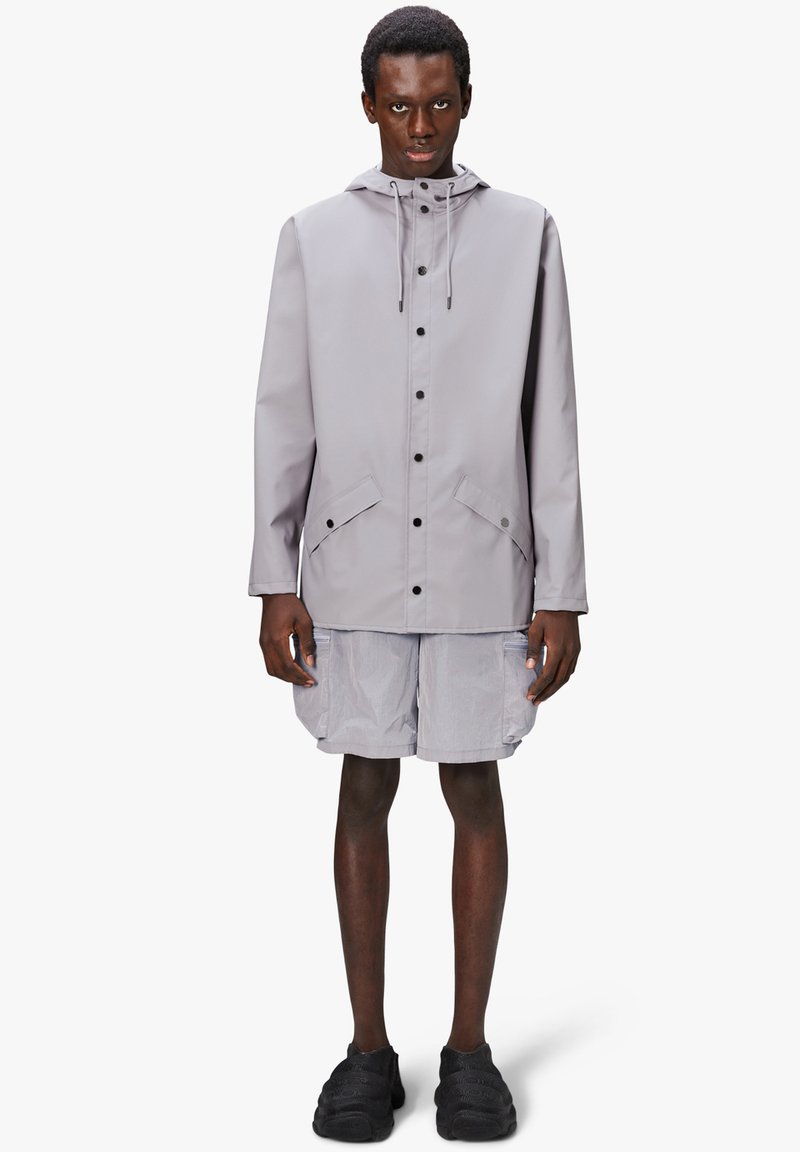 Jasnoszara kurtka wodoodporna z kapturem, na zatrzaski, z dwoma przednimi kieszeniami, noszona na jasnoszare szorty i czarne buty.