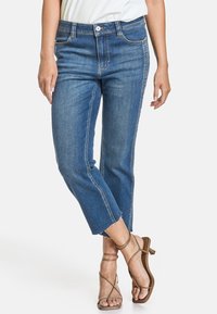 Žena v mid-rise modrých džínách s zkrácenými, roztrhanými lemy a hnědými sandály na podpatku, stojící se zkříženýma nohama.