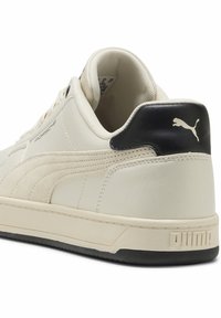 Puma fodtøj i cremefarve med sorte detaljer, der har en blød læderoverdel, tekstureret midtersektion og en gummisål med et logo på hælen.