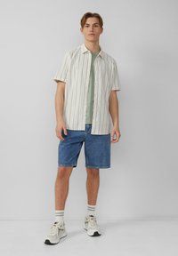 Koszulka z krótkim rękawem w kremowe paski na zielonej koszulce, połączona z niebieskimi szortami dżinsowymi. Sportowe buty z jasnymi akcentami dopełniają outfit.