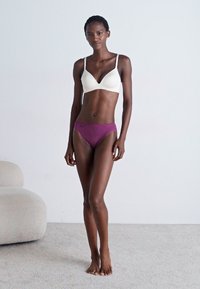 Brassière blanche et culotte hipster violette, design sans coutures, tissu doux, style minimaliste, forme ajustée, portée par un modèle en position debout.