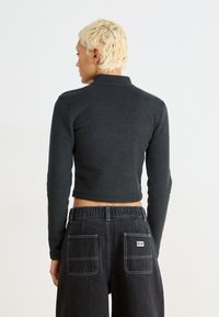 Czarny, krótki polarowy top z długimi rękawami i wysokim kołnierzem, noszony z ciemnymi dżinsami z wysokim stanem, posiadającymi dwie tylne kieszenie i białe przeszycia.