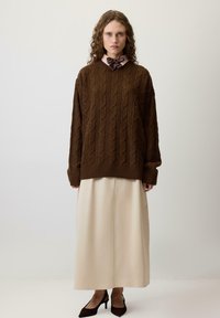 Brown cable-knit sweater with oversized fit, layered over a cream midi skirt. Features a high neckline and extended sleeves. Black slingback heels.