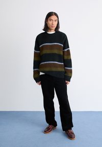 Chunky strikket sweater i mørkegrøn med brun, oliven og lyseblå stribe-mønster. Kombineret med sorte fløjl-bukser og brune sko.