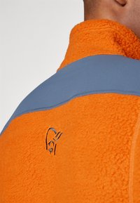 Fleece jacket with an orange body and blue shoulder panels, featuring a textured orange surface and a logo embroidered near the neck.
