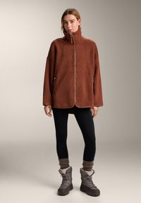 Brown fleece jacket with high collar and zip front, loose fit, two side pockets, paired with black leggings and gray winter boots.