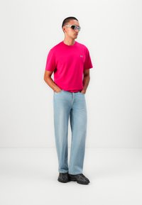 Lys pink T-shirt med et lille logo, lyseblå brede jeans og sorte teksturerede sneakers. Modellen bærer grå solbriller.