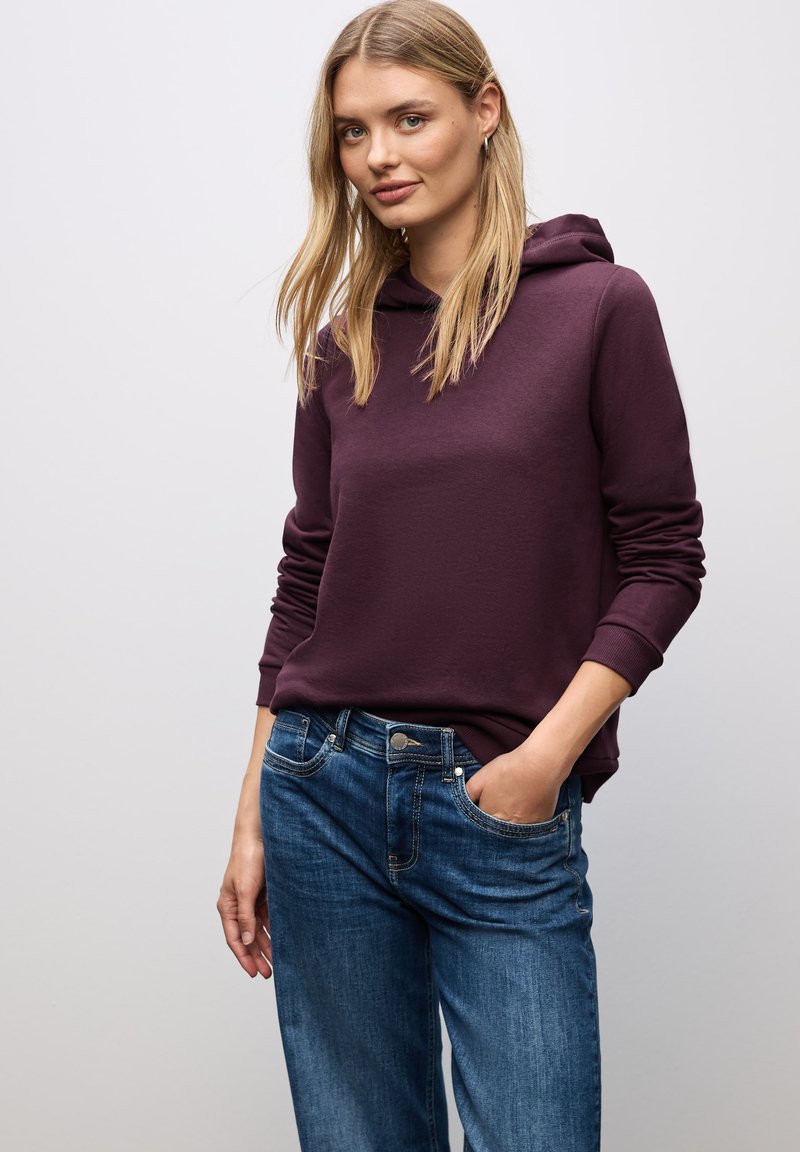 Sudadera burdeos con una textura suave y bolsillo frontal, combinada con pantalones vaqueros azules. Las mangas están ligeramente recolectadas en los puños.