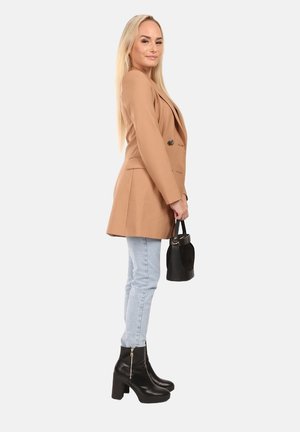 Beige dubbelzijdig blazer met een getailleerde pasvorm, gecombineerd met lichtblauwe skinny jeans en zwarte enkellaarsjes met hak. Houdt een kleine zwarte tas vast.