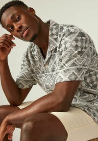 Camisa de manga corta en un tejido texturizado con un patrón geométrico en blanco y negro. Presenta un cuello y botones marrones. Se llevan unos pantalones cortos beige.