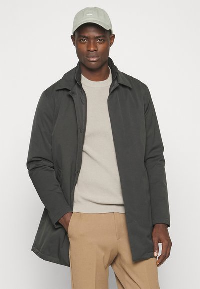 Abrigo gris oscuro con cierre de botones, cuello de camisa y mangas largas; llevado sobre un suéter beige claro con textura acanalada, complementado con pantalones caqui.