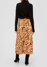 Kvinna iklädd en svart långärmad topp, plisserad midikjol med orange, svart och beige abstrakt mönster, samt svarta klackboots, stående med ryggen vänd.