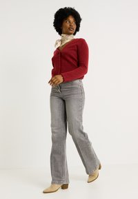 Cárdigan de lana roja con botones en la parte delantera, vaqueros acampanados de talle alto grises y botas de tacón beige. Accesorios con una bufanda de patrón claro.