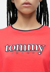 Közeli kép egy piros inget viselő személyről, amelynek mellkasán fehér és tengerészkék "tommy JEANS" hímzés található.
