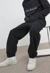 Person sitting on a metal-framed chair wearing black Karl Kani tracksuit and white sneakers on beige carpet against a plain white wall.