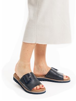 Feet wearing black slide sandals with large buckles and brown soles, standing on a white floor with beige trousers visible.