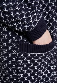 Virkat marinblå tröja med vit invecklad mönster. Har ribbade manschetter och en ficka på sidan. Strukturerad tyg med synliga stygn.