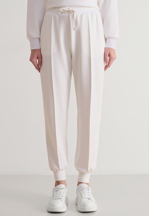 Pantaloni da jogging bianchi con una vestibilità rilassata, dotati di vita con cordino, polsini a costine e pieghe frontali. Indossati con sneakers bianche.