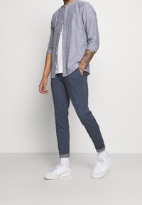 Svetle modrá košile na knoflíky, bílý tričko pod ní, pruhované námořnické kalhoty a bílé tenisky. Má ohrnuté rukávy a slim fit střih.