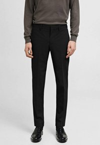 Pantalons noirs ajustés avec une texture lisse, des détails de pli avant et une coupe classique, associés à une chemise noire à manches longues et des chaussures noires.